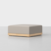 A modern, square ottoman with a light gray upholstered cushion and a natural wood base, placed on a smooth, light-colored floor against a plain white background.