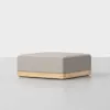 A modern, square ottoman with a light gray upholstered cushion and a natural wood base, placed on a smooth, light-colored floor against a plain white background.