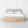 A modern bed with a light wood frame, a gray upholstered headboard, and white bedding with two pillows, set against a plain white wall in a minimalistic room.