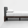 A side view of a neatly made bed with a dark wooden frame, a gray upholstered headboard, white sheets, and a single white pillow against a plain white wall.