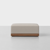 A modern rectangular ottoman with light beige upholstery and a wooden base, placed on a light-colored floor against a plain white background.