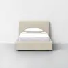 A beige upholstered bed with a white mattress, white sheets, and a white pillow, positioned against a plain white wall in a minimalistic room with a gray floor.