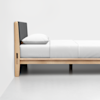 A modern wooden bed frame with a dark upholstered headboard, white pillow, and white bedding, positioned against a plain white wall and floor.