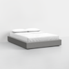 A minimalist grey platform bed with a low profile, dressed in white bedding and two white pillows, set against a plain white wall and floor in a modern, empty room.