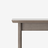 A close-up of the corner of a light gray wooden table, showing the tabletop, leg, and apron against a plain white background.