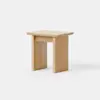 A small, light wood stool with a flat rectangular top and two wide, sturdy legs. The design is simple and modern, with clean lines and a natural finish. The stool is placed on a white background.