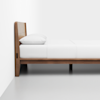 A wooden bed frame with a white mattress, white sheets, and a white pillow is positioned against a white wall on a light gray floor. The image is taken from the side, showing the minimalist design.