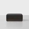 A modern, rectangular ottoman with a dark fabric cushion and a wooden base, placed on a light grey surface against a plain white background.