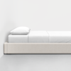A minimalist side view of a bed with a beige upholstered frame, neatly made with white sheets and a single white pillow, placed against a plain white wall on a smooth, light gray floor.