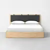 A minimalist wooden bed with a rectangular frame and a black cushioned headboard. The bed is neatly made with white bedding and two pillows, set against a plain white wall on a gray floor.