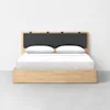 A minimalist wooden bed with a rectangular frame and a black cushioned headboard. The bed is neatly made with white bedding and two pillows, set against a plain white wall on a gray floor.