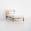 A minimalist wooden single bed with a headboard, white bedding, and a single pillow, placed in a plain, light-colored room.
