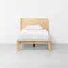 A simple wooden bed frame with a headboard, dressed with a white pillow and white bedding, stands on a light gray floor against a plain white wall.