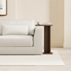 A modern living room features a light-colored sofa with a rectangular pillow. Beside it is a wooden side table holding a cup and stacked books. The room has neutral walls and a light rug.