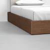 A close-up of a wooden platform bed with a white mattress, white bedding, and a light-colored headboard, placed on a light gray floor.