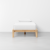 A simple wooden bed frame with a mattress and pillow, all covered in white bedding, stands against a plain white wall on a smooth light gray floor.