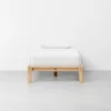 A simple wooden bed frame with a mattress and pillow, all covered in white bedding, stands against a plain white wall on a smooth light gray floor.