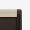 Close-up of the corner of a dark wooden bed frame with a light-colored, cushioned headboard panel partially visible, against a plain white background.