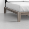 Close-up of a wooden bed frame with light brown legs and a neatly made bed with white sheets in a minimal, modern room with a light gray floor and white walls.