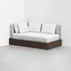 A modern daybed with a dark wooden frame, light beige upholstered backrest, and four white pillows, placed in the corner of a minimalist room with white walls and a light gray floor.