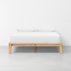 A minimalist wooden bed frame with a light wood finish, neatly made with white bedding and two white pillows, set against a plain white wall in a modern, clean room.