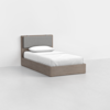 A single bed with a wooden frame and a padded gray headboard, dressed in white bedding and a pillow, placed in a minimal, empty room with light walls and floor.