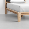 A close-up view of a modern wooden bed frame with clean lines and light-colored wood, supporting a neatly made bed with white sheets and a pillow, on a smooth gray floor.