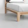 A close-up view of a modern wooden bed frame with clean lines and light-colored wood, supporting a neatly made bed with white sheets and a pillow, on a smooth gray floor.