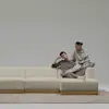 A man and a woman in casual, neutral-toned outfits relax on a minimalist cream-colored sectional sofa with a brass base, set against a plain, light gray background.