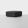 A large, square, dark gray ottoman with a minimalist design sits on a light gray floor against a plain white background.