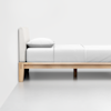 Minimalist bed with a light wooden frame, white mattress, white bedding, and a single white pillow, placed against a plain white wall on a smooth, light-colored floor.
