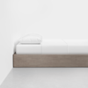 A minimalistic bed with a wooden frame, white mattress, white sheet, and a single white pillow, positioned against a plain white wall on a light grey floor.
