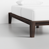Close-up of a modern wooden bed frame with dark brown legs, supporting a neatly made bed with white sheets and a white pillow, against a plain white wall and light floor.