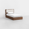 A single bed with a wooden frame and a padded, light-colored headboard is placed in a minimalist room with white walls and a light gray floor. The bed is made with white linens and one pillow.