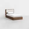 A single bed with a wooden frame and a padded, light-colored headboard is placed in a minimalist room with white walls and a light gray floor. The bed is made with white linens and one pillow.