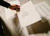 A hand holds up a technical drawing of a mechanical or architectural structure over a table covered with other similar technical sketches and a cutting mat.