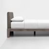 A minimalist bed with a wooden frame and legs, topped with a white mattress, white bedding, and a single white pillow, placed against a plain white wall in a bright room.