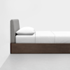 A minimalist bed with a wooden frame, white bedding, a single pillow, and a gray upholstered headboard, positioned against a white wall on a light gray floor.