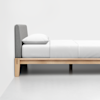 A minimalist wooden bed frame with light wood legs, a gray upholstered headboard, and neatly arranged white bedding with a pillow, set against a plain white wall and floor.