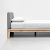 A minimalist wooden bed frame with light wood legs, a gray upholstered headboard, and neatly arranged white bedding with a pillow, set against a plain white wall and floor.