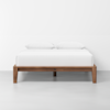 A minimalist wooden bed frame with a white mattress and two white pillows, set against a plain white wall in a bright, uncluttered room with a light gray floor.