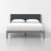 A modern bed with a gray upholstered headboard, light wood frame, white bedding, and two white pillows, set against a plain white wall and floor.