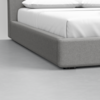 A close-up view of the corner of a modern bed with a gray upholstered frame, white bedding, and pillows in a minimalist room with a light gray floor.