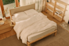 A cozy, neatly made bed with white bedding is in a sunlit wooden bedroom. There is a wooden nightstand with a vase, an empty shelving unit, and large windows with sheer curtains letting in natural light.