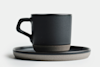 A matte black ceramic coffee cup with a handle sits on a matching saucer. Both have a minimalist design with a subtle brown base, set against a plain white background.