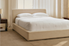 A neatly made bed with a beige upholstered frame and headboard, white bedding, and two white pillows, sits in a softly lit bedroom with cream walls, large windows, and minimal wooden furniture.
