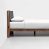 A minimalist wooden bed frame with a dark upholstered headboard, white bedding, and pillow, shown from the side against a plain white wall and floor.