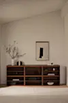 A minimalist living room with light walls features a dark wooden console holding books, pottery, and decor. A white vase with green branches and abstract wall art add subtle accents. The space feels calm and uncluttered.