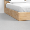 A close-up view of the corner of a light wood bed frame with white bedding and pillow, set on a light gray concrete floor.