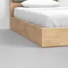 A close-up view of the corner of a light wood bed frame with white bedding and pillow, set on a light gray concrete floor.